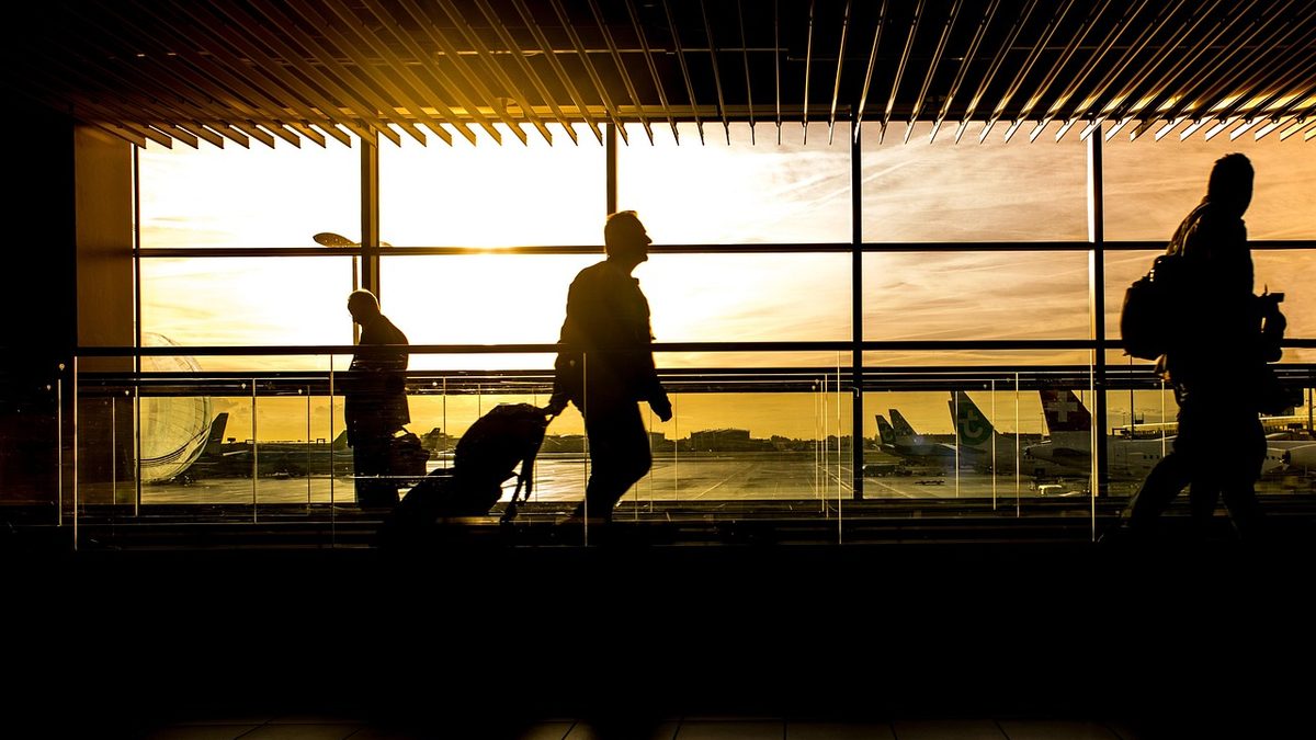 Reisende mit Gepäck vor der Fensterfront eines Flugsteigs, dahinter Flugzeuge im Abendlicht