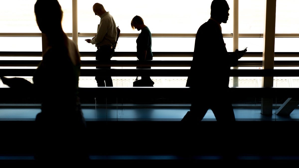 Silhouetten mehrerer Reisender, die hintereinander an einer hellen Fensterfront entlanggehen