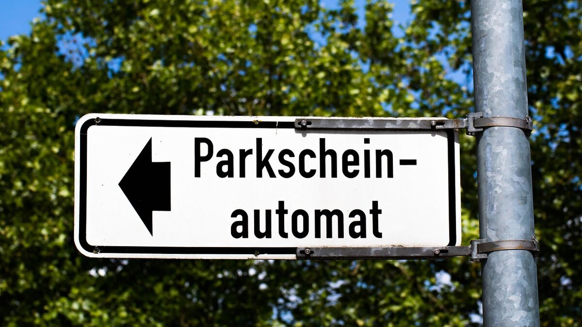 Wegweiser mit der Aufschrift „Parkscheinautomat" und einem Richtungspfeil, montiert an einem Mast vor Laubbäumen